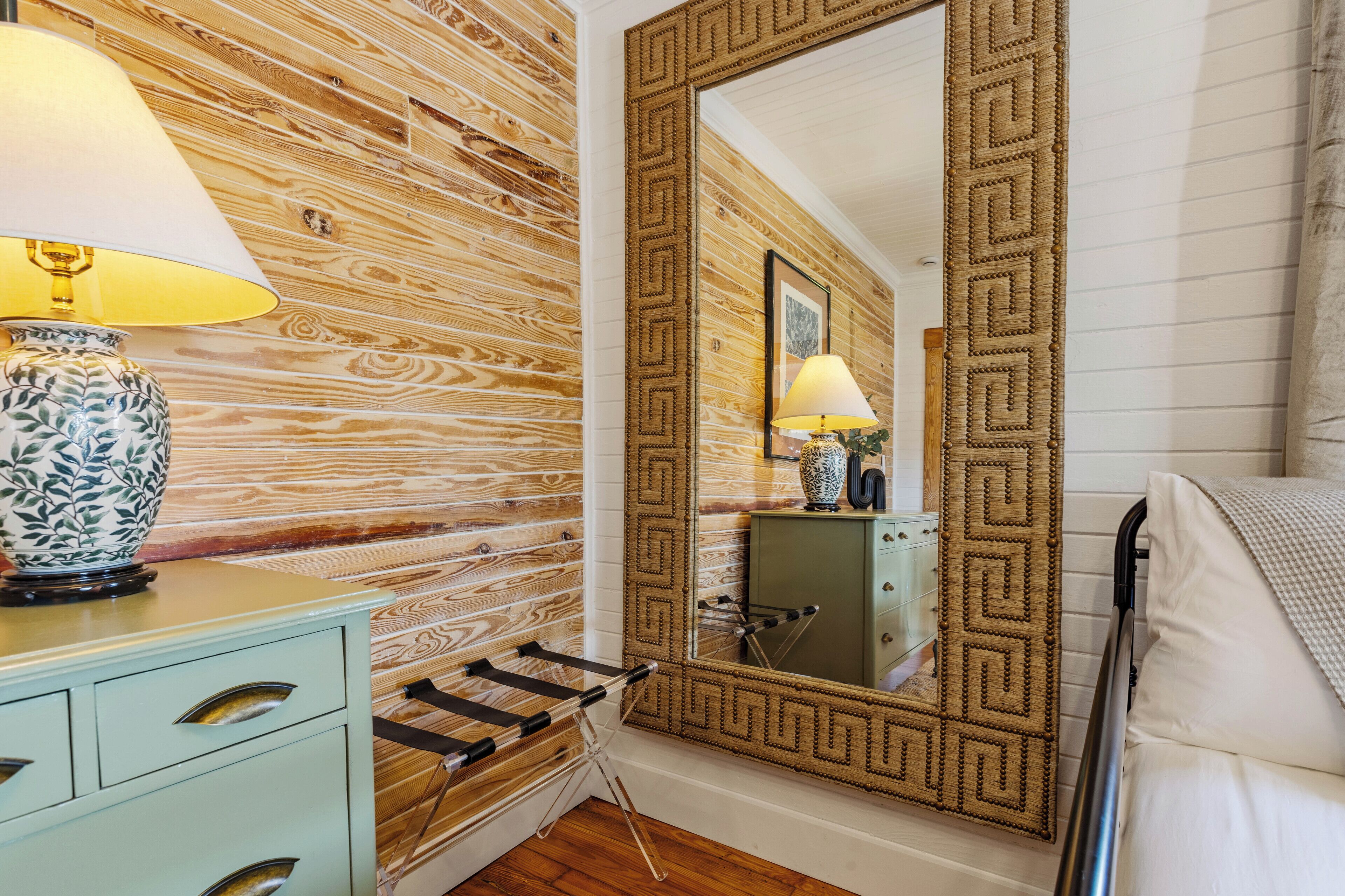 A room with a wooden wall, a large mirror with a geometric frame, a bedside table with a lamp, and a bed with a blanket.