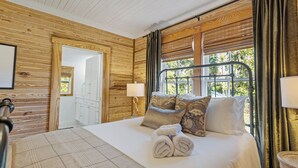 A wooden-paneled room with a bed, a window with curtains, and a door leading to a bathroom. - Welcome to The Schoolhouse – A charming cottage in historic Monticello, Florida. (Monticello)