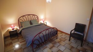 A bedroom with a metal bed frame, a single bedside table with a lamp, a chair, and a hanging light fixture.