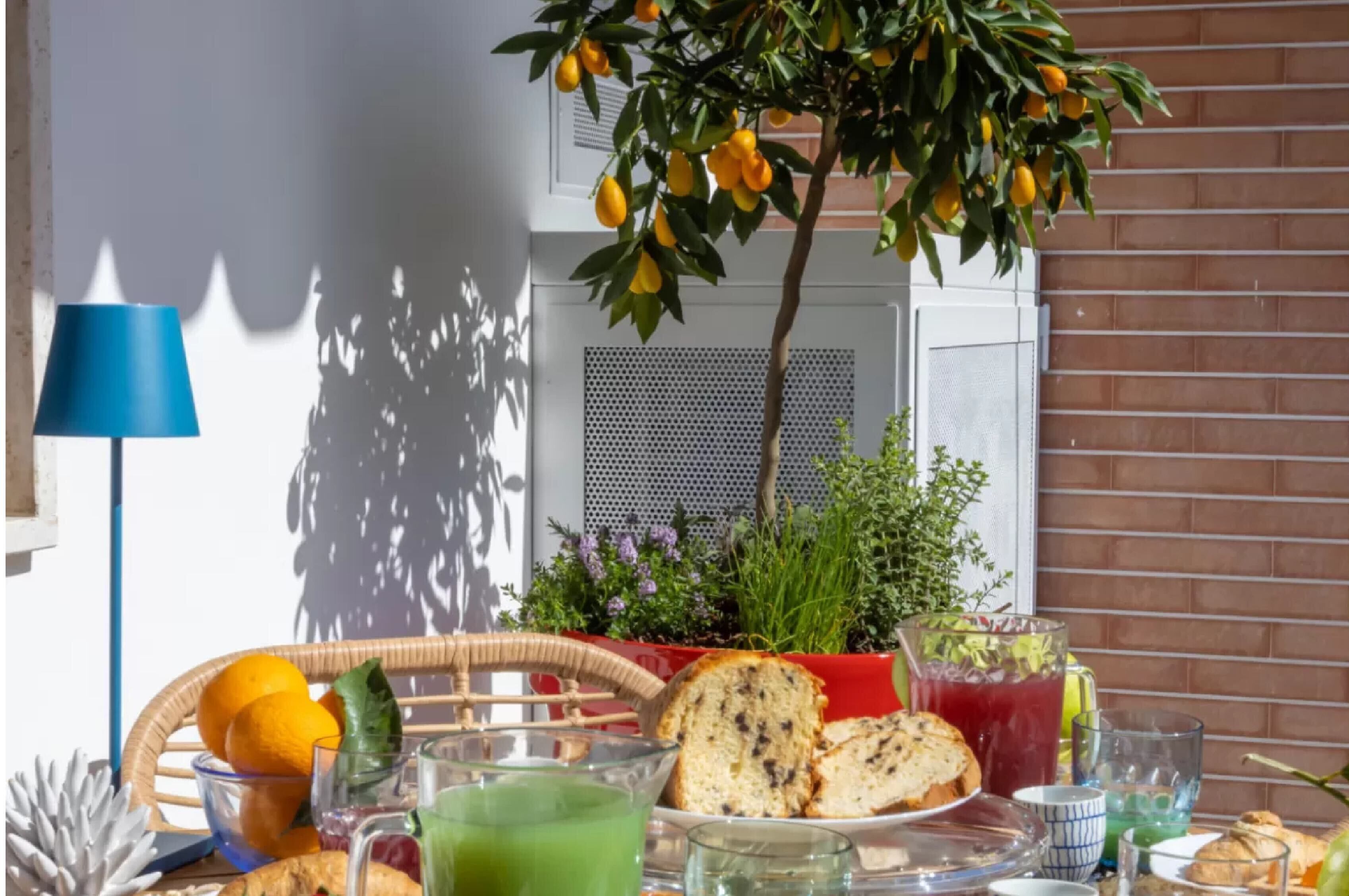 A breakfast table with fruits, bread, and drinks, a wicker chair, a potted plant, and a blue lamp.