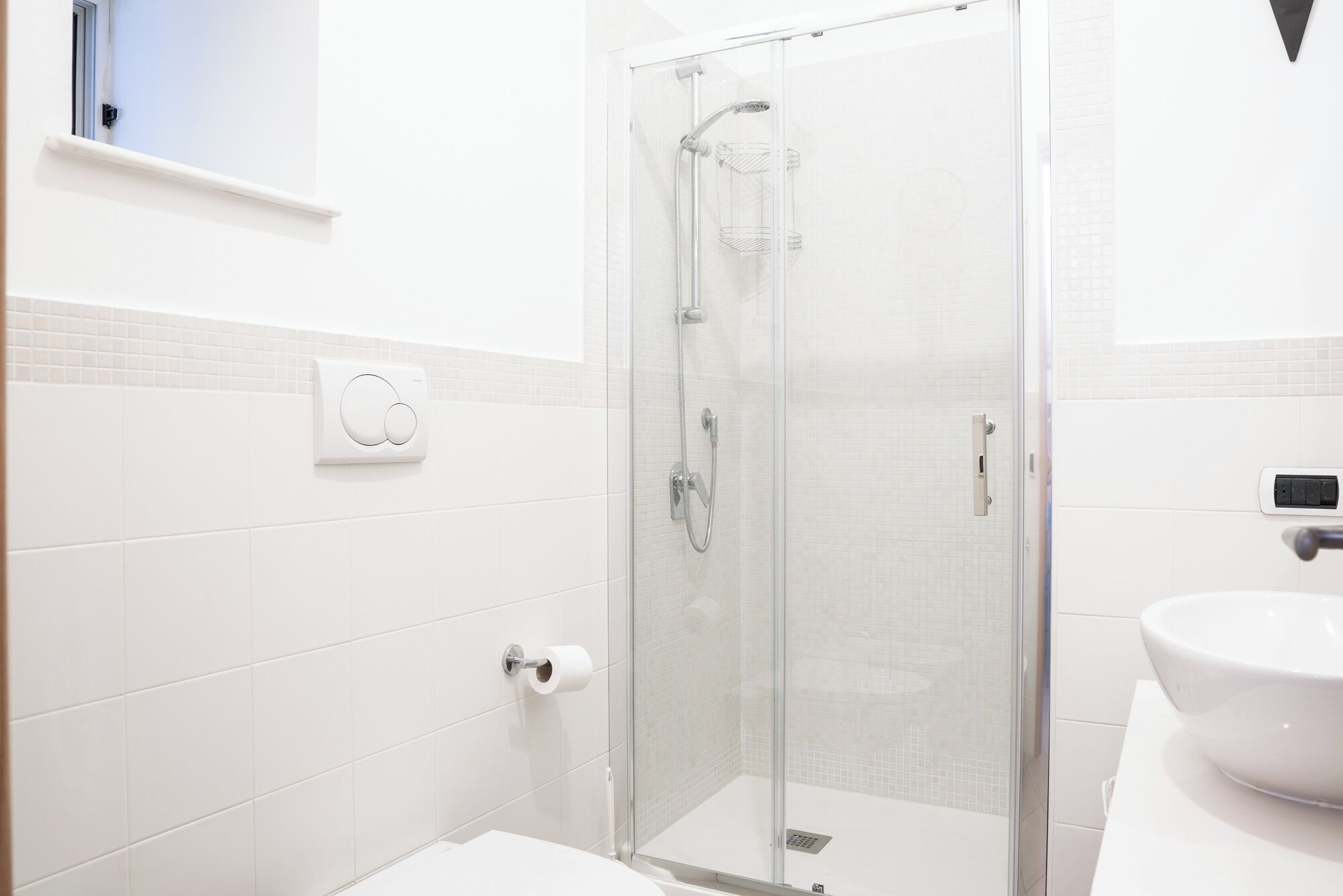 A modern bathroom with a glass-enclosed shower, white tiles, a toilet paper holder, and a sink.