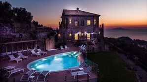 A stone building with a pool and lounge chairs, surrounded by a stone wall and a view of the sea at sunset. - Villa Breathtaking View Near Nerano (Massa Lubrense)