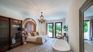 A spacious bedroom with a large bed, a sitting area with a chair, a bathtub, and a view of greenery through the windows. - Villa Chianti With Infinity Pool And Vineyard View (Greve in Chianti)
