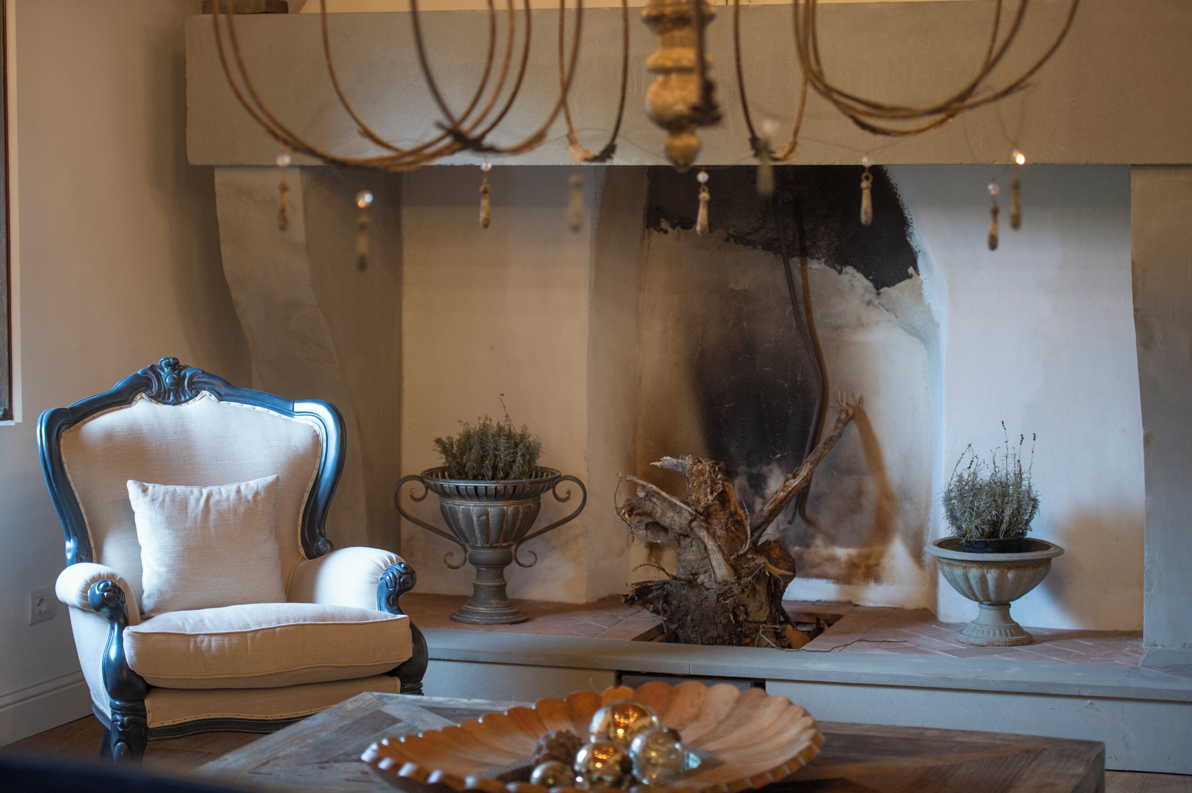 Um quarto com uma poltrona clássica, duas urnas decorativas com plantas, uma escultura em madeira e um candelabro.