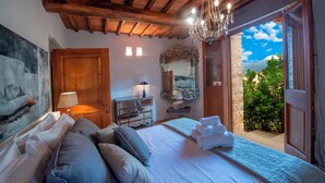 Une chambre avec un grand lit, un plafond en bois, un lustre, un mur en pierre et une vue sur la verdure.