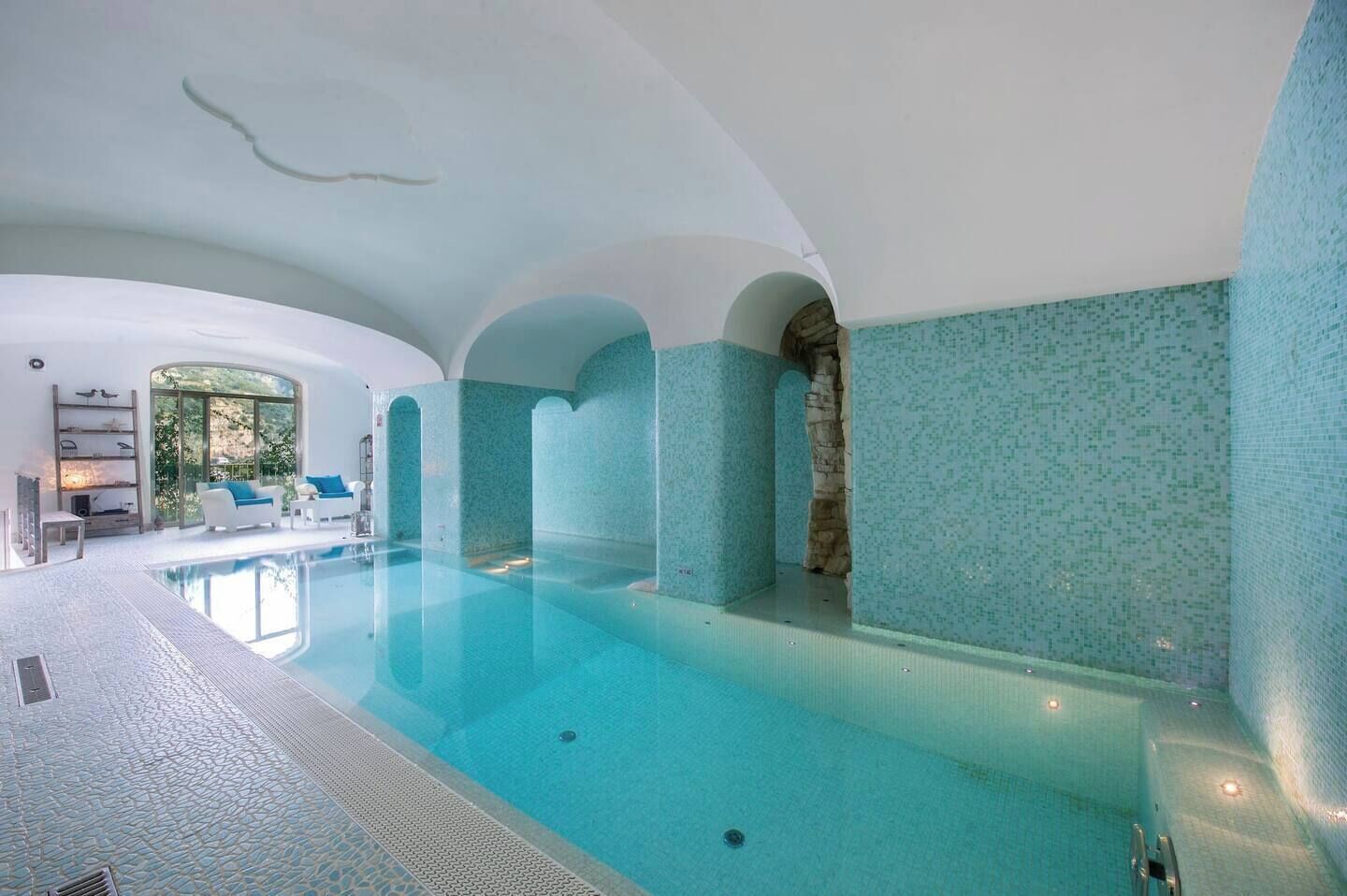 A large indoor swimming pool with a tiled floor and walls, surrounded by white arches and a seating area with blue cushions.