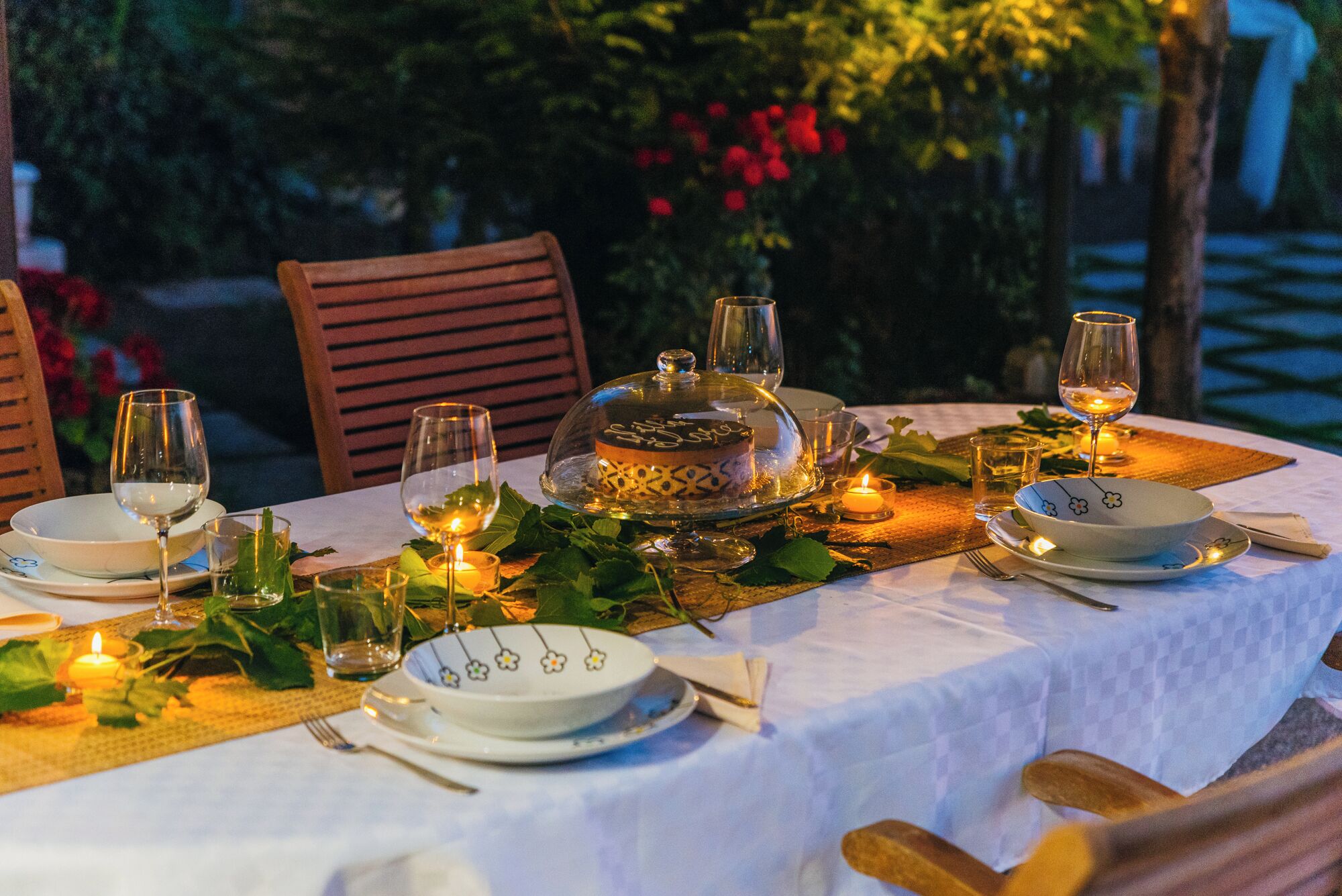 A table set for a meal with a dessert on a glass stand, surrounded by candles and greenery.