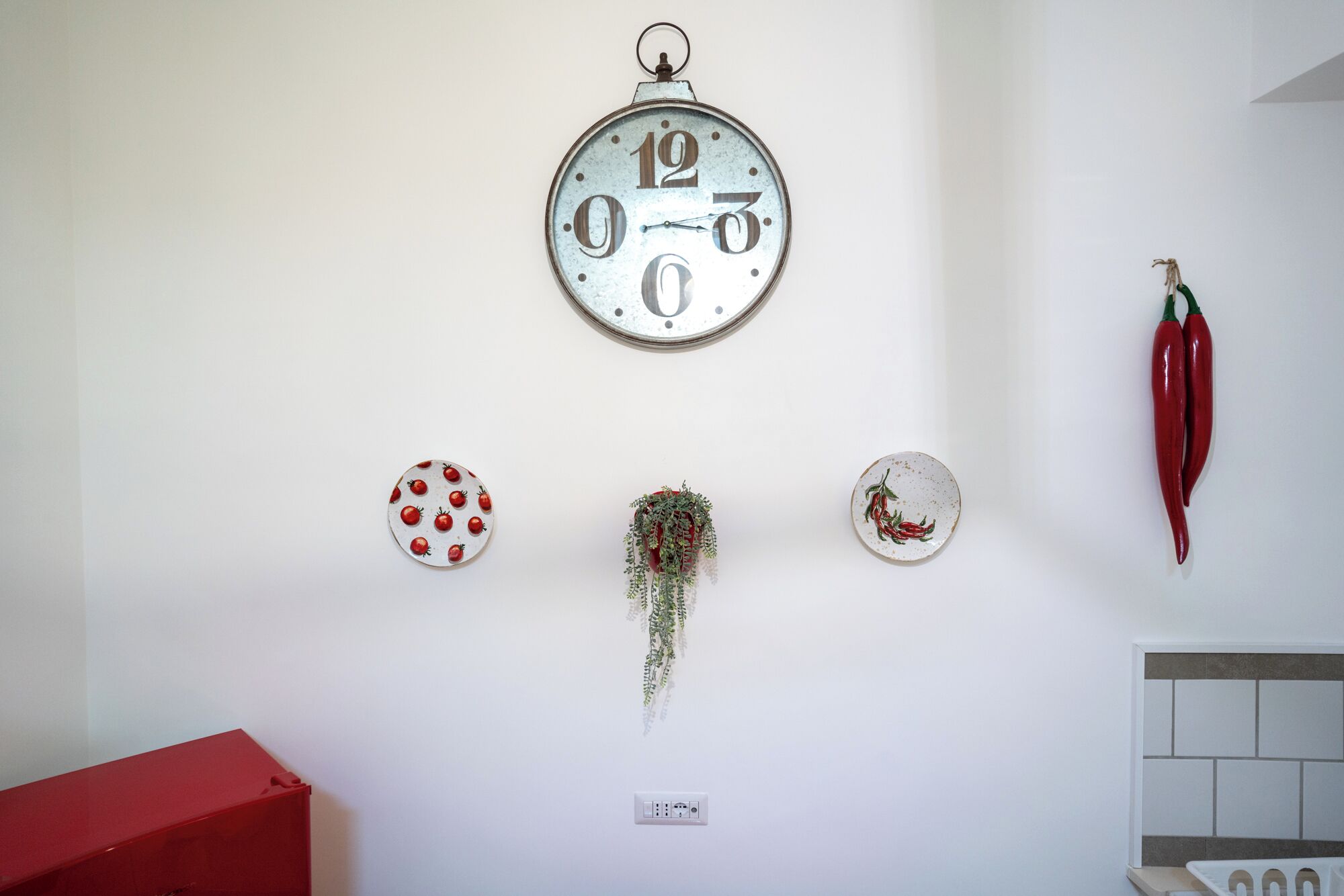 A wall with a clock, a red box, a power outlet, and decorative items including chili peppers and a plant.