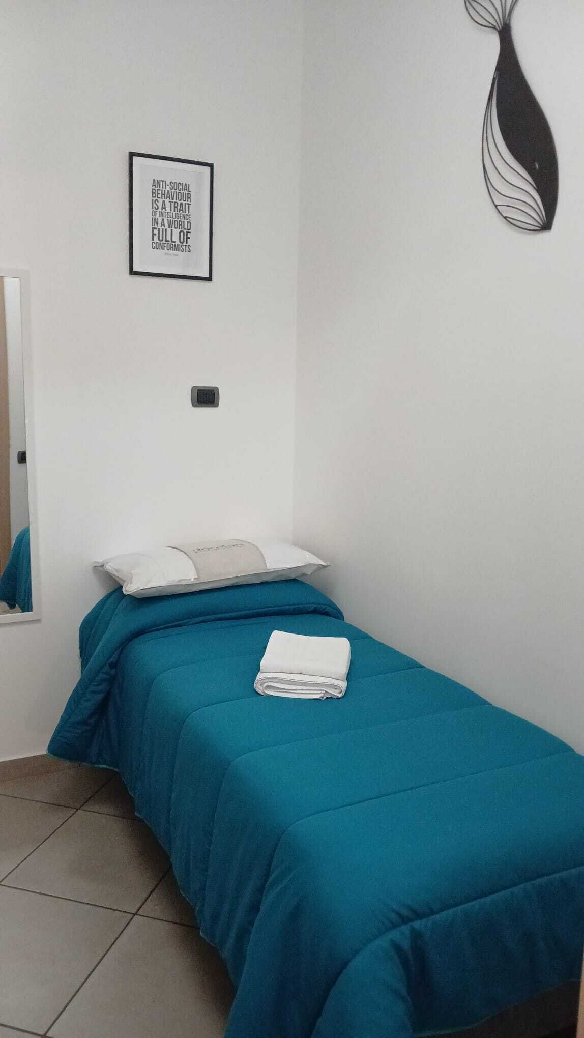 Une chambre avec une couvre-lit turquoise, un oreiller blanc, une serviette blanche pliée, un luminaire fixé au mur et une citation encadrée accrochée au mur.