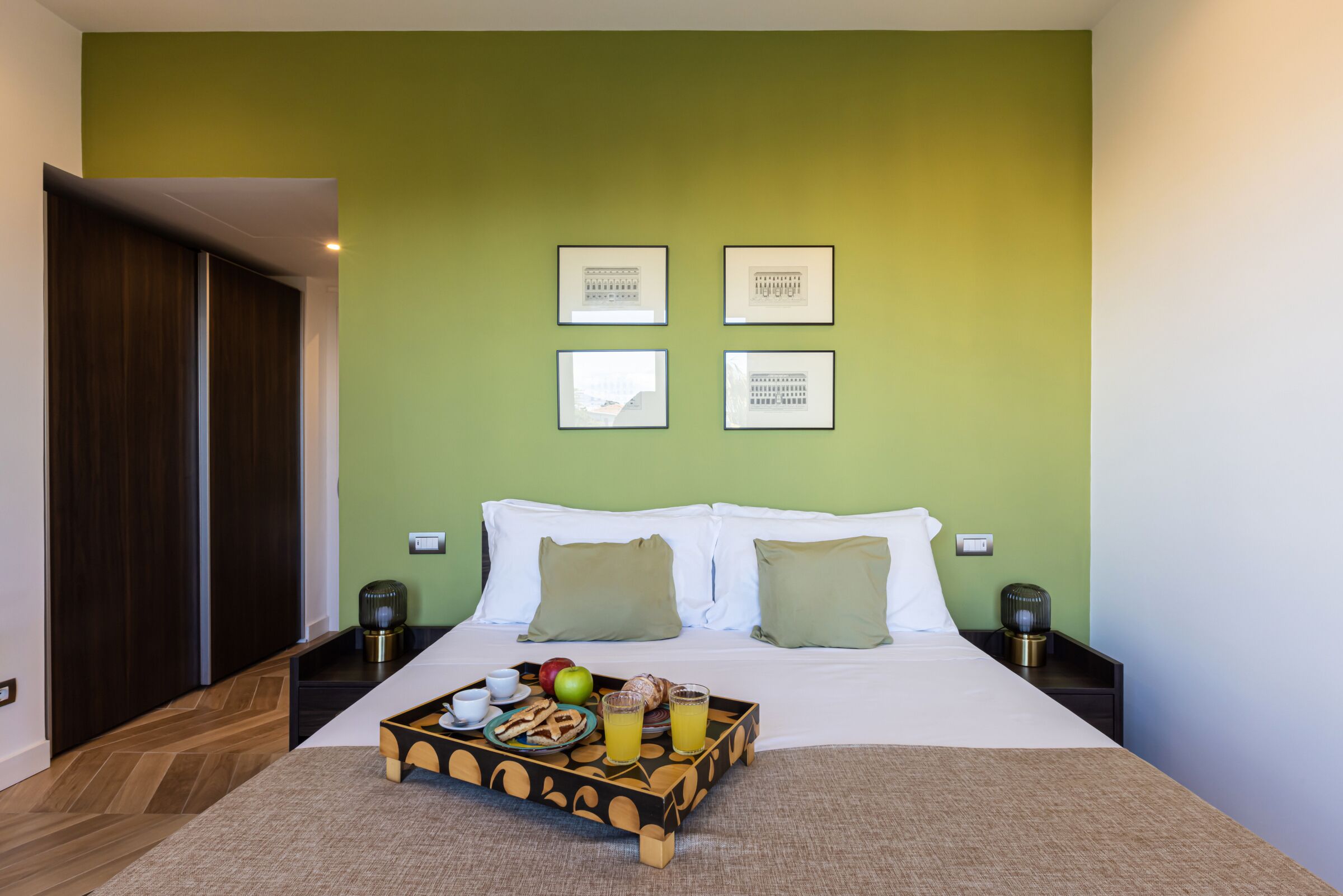 Una cama bien tendida con ropa blanca, una bandeja con alimentos para el desayuno, incluyendo frutas y pasteles, y una pared verde con cuadros.