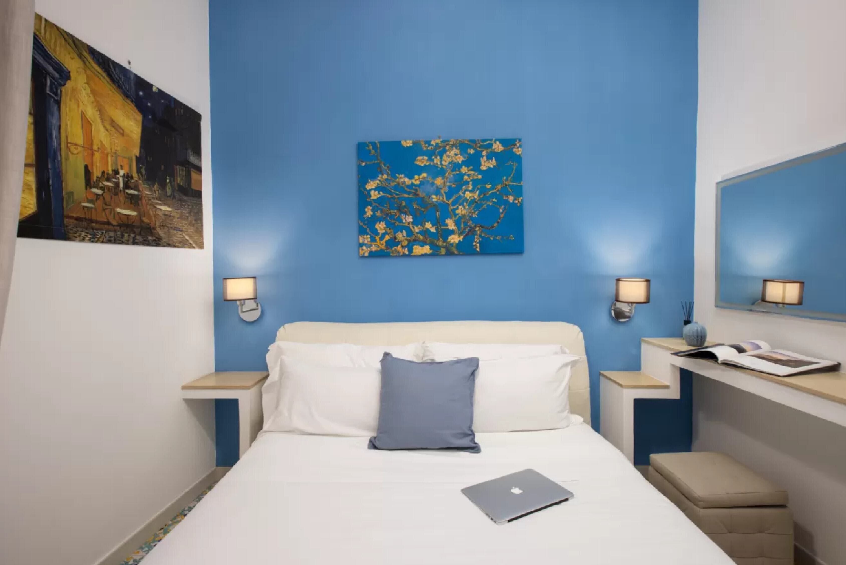 Habitación de hotel con cama, una computadora portátil, un cuadro abstracto azul y una pintura de escena nocturna.