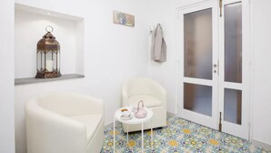 A small, white-walled room with a white armchair, a small round table with a teapot and cups, a decorative lantern, and a door with frosted glass panels. - Brezza Marina-Authentic Sorrento Flat By The Beach (Sorrento)