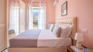 Une chambre à coucher avec un lit, une table de chevet, une lampe, un tableau encadré et une vue sur les bâtiments à travers des rideaux transparents.