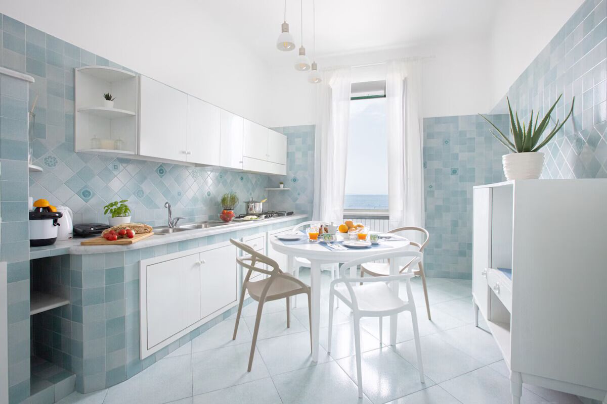 Une cuisine moderne avec des meubles blancs, une table à manger avec des chaises et une vue sur la mer par une fenêtre.