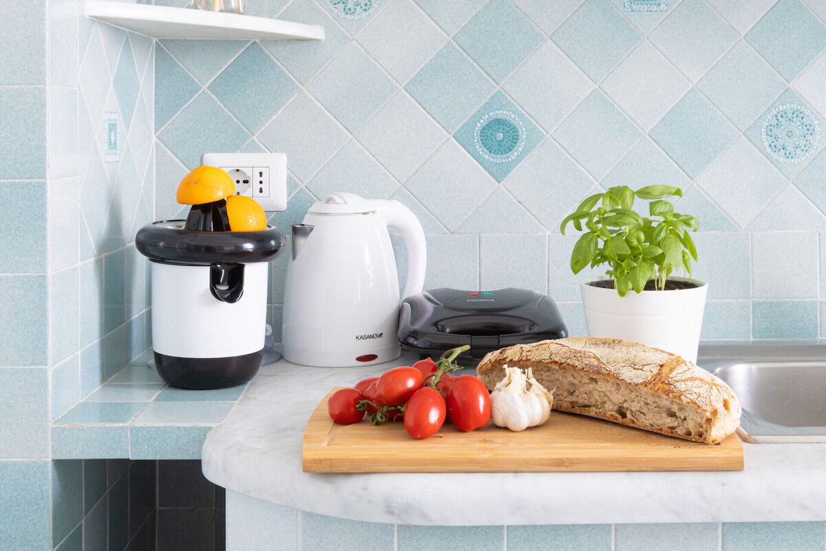A kitchen counter with a bread loaf, tomatoes, garlic, a juicer, a kettle, and a potted basil plant.