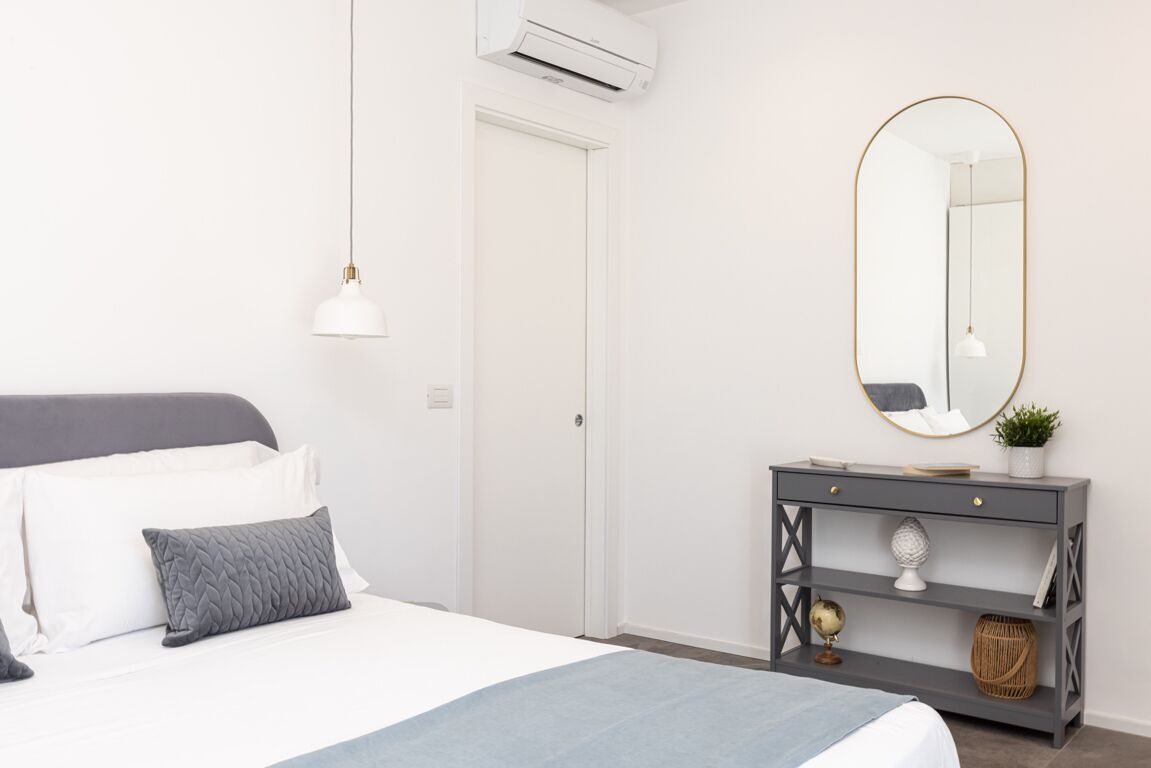 A bedroom with a bed, bedside table, mirror, and an air conditioning unit.