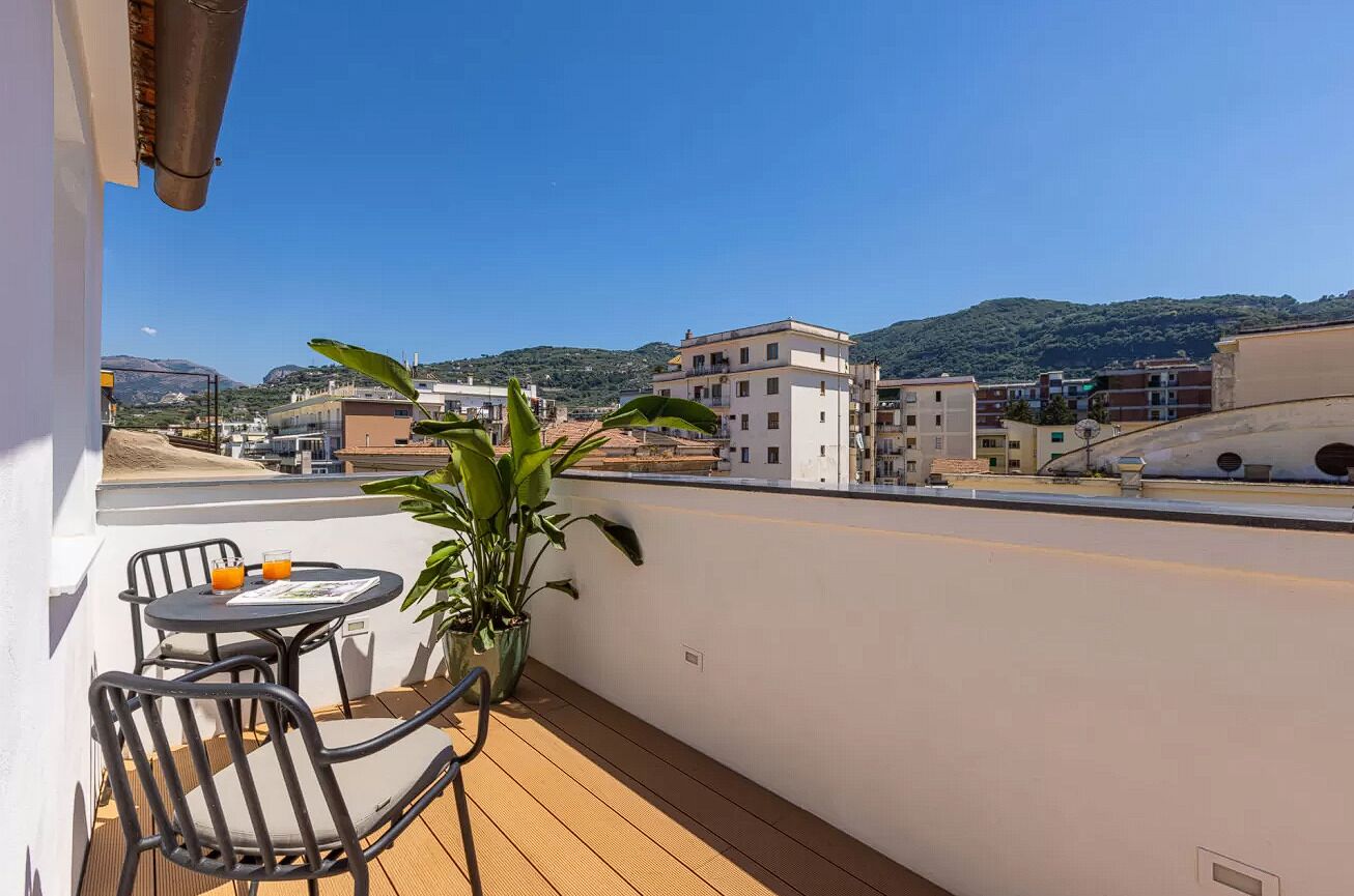 A balcony with a table and chairs, a potted plant, and a view of buildings and hills in the background.