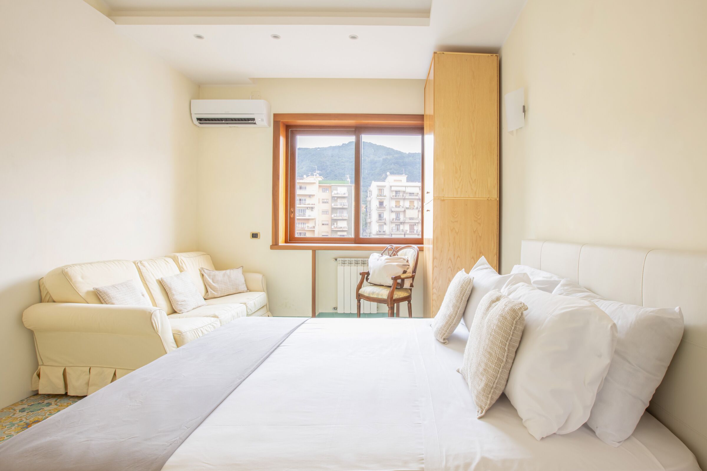 Un dormitorio con cama, sofá, sillón y ventana con vistas a edificios y montañas.