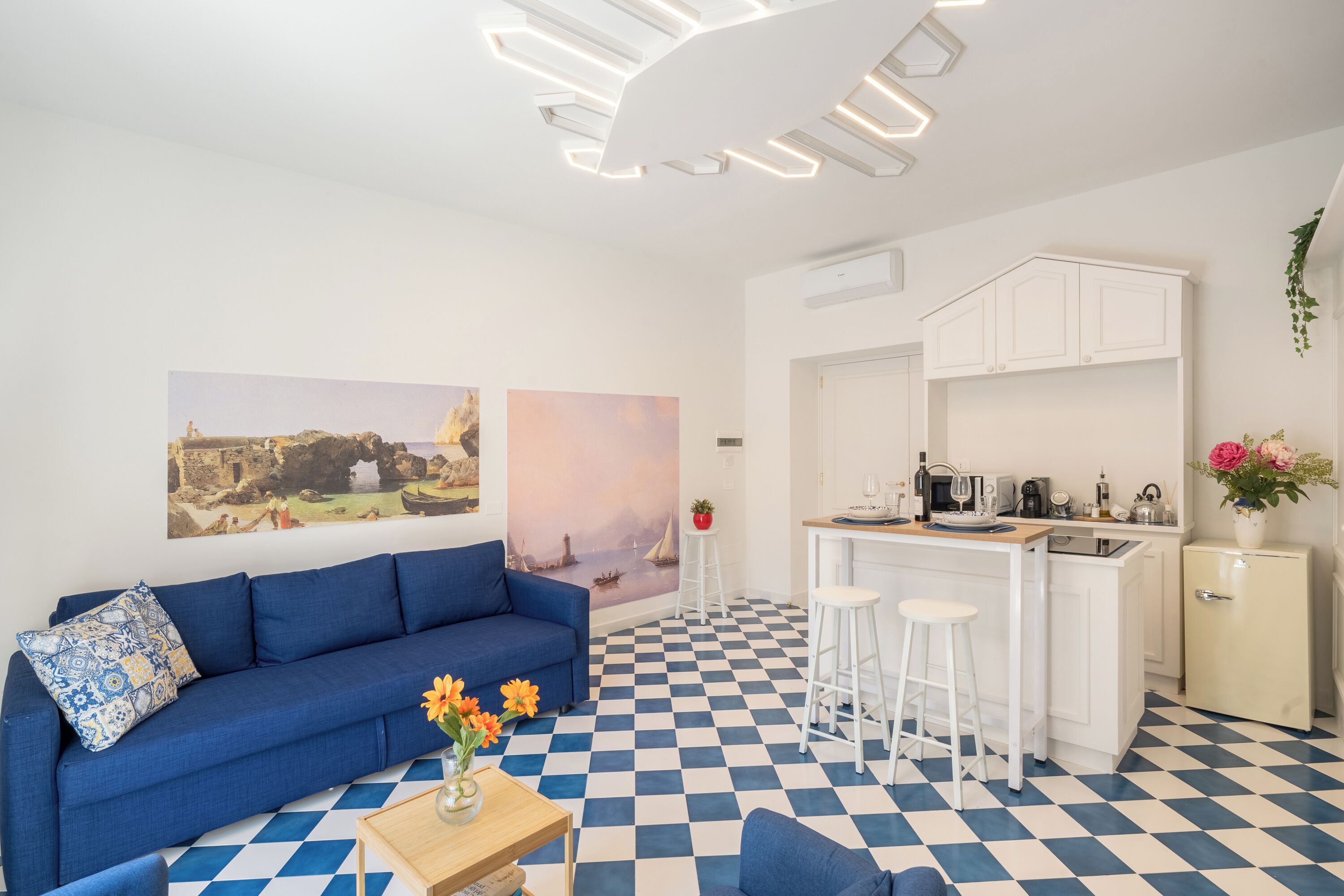 Uma sala de estar moderna com um sofá azul, piso xadrez e uma área de cozinha com armários brancos e um banquinho de bar.