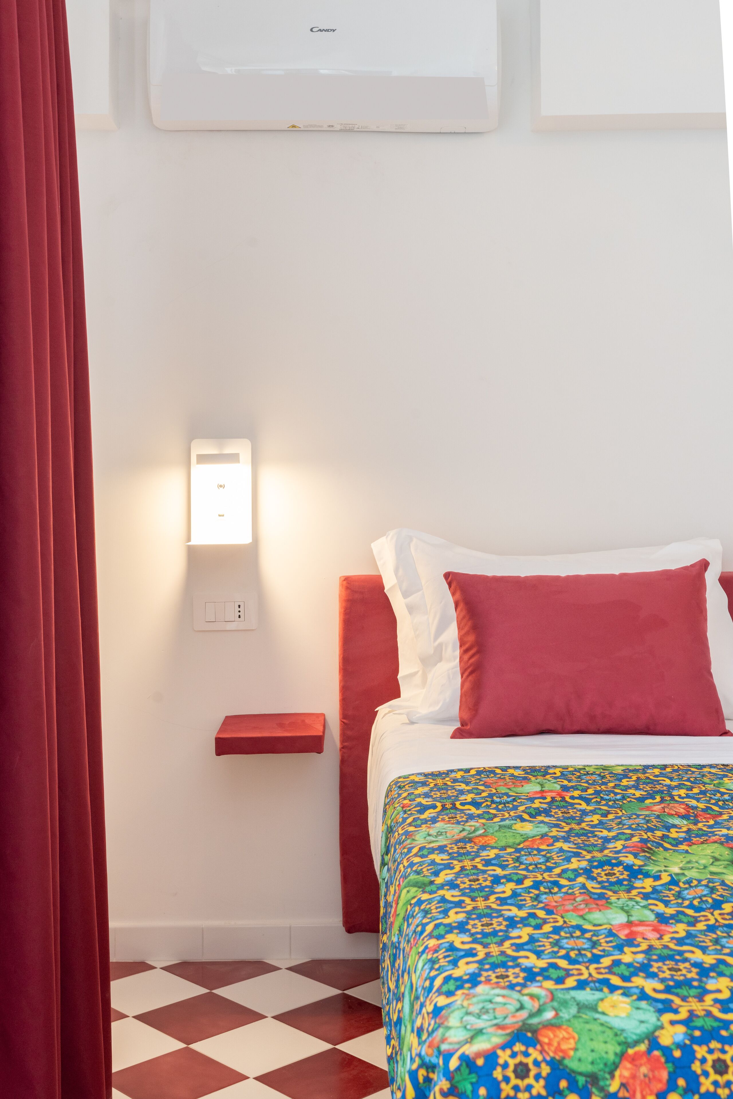 Une chambre d’hôtel avec un lit, une table de chevet, un rideau rouge, une couvre-lit à motifs et un sol à carreaux.