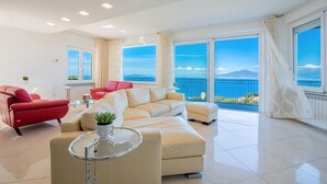 A modern living room with a beige sectional sofa, a red armchair, and a glass coffee table. Large windows offer a view of the sea and a distant mountain. - Villa With Panoramic Vesuvius And Sorrento Views With Private Pool (Sorrento)