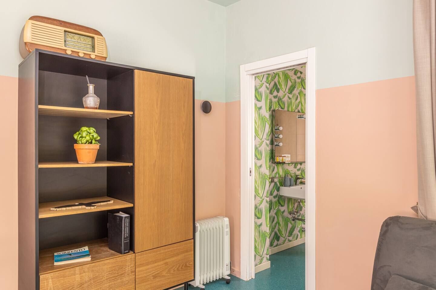 A room with a wooden wardrobe, a potted plant, a radiator, and a bathroom with a leaf-patterned wallpaper.