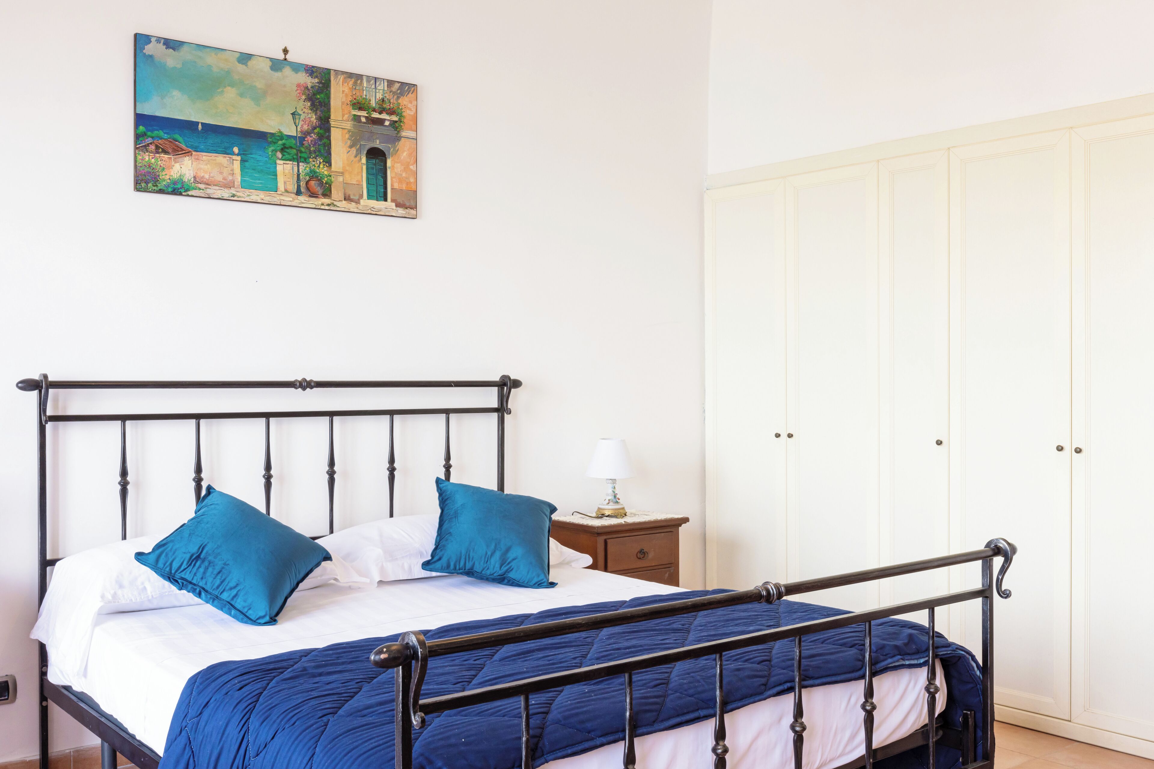 Une chambre à coucher avec un lit, des oreillers, une table de chevet, une lampe et un tableau représentant un paysage côtier.