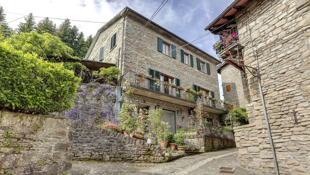 A stone building with a balcony adorned with flowers, a stone pathway, and lush greenery.
