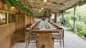 A long wooden dining table set with chairs, surrounded by lush greenery and a thatched roof.