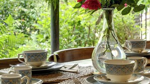 A table set with blue and white patterned cups and saucers, a clear glass vase with red flowers, and a view of lush greenery and a distant building.
