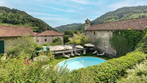 A pool area with lounge chairs and umbrellas, surrounded by lush greenery and stone buildings with a hill in the background.