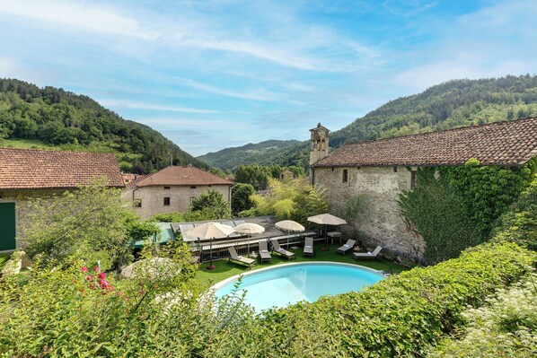A pool area with lounge chairs and umbrellas, surrounded by lush greenery and stone buildings with a hill in the background. - Entire Hilltop Villa With Private Pool In Palazzuolo Sul Senio (Palazzuolo sul Senio)