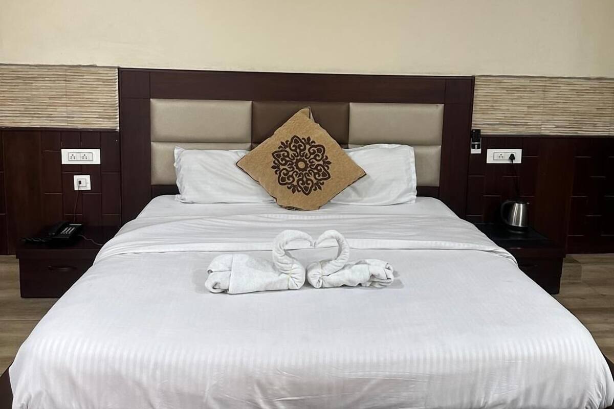 A neatly made bed with a heart-shaped towel arrangement, a decorative pillow, and a bedside table with a phone and a kettle.