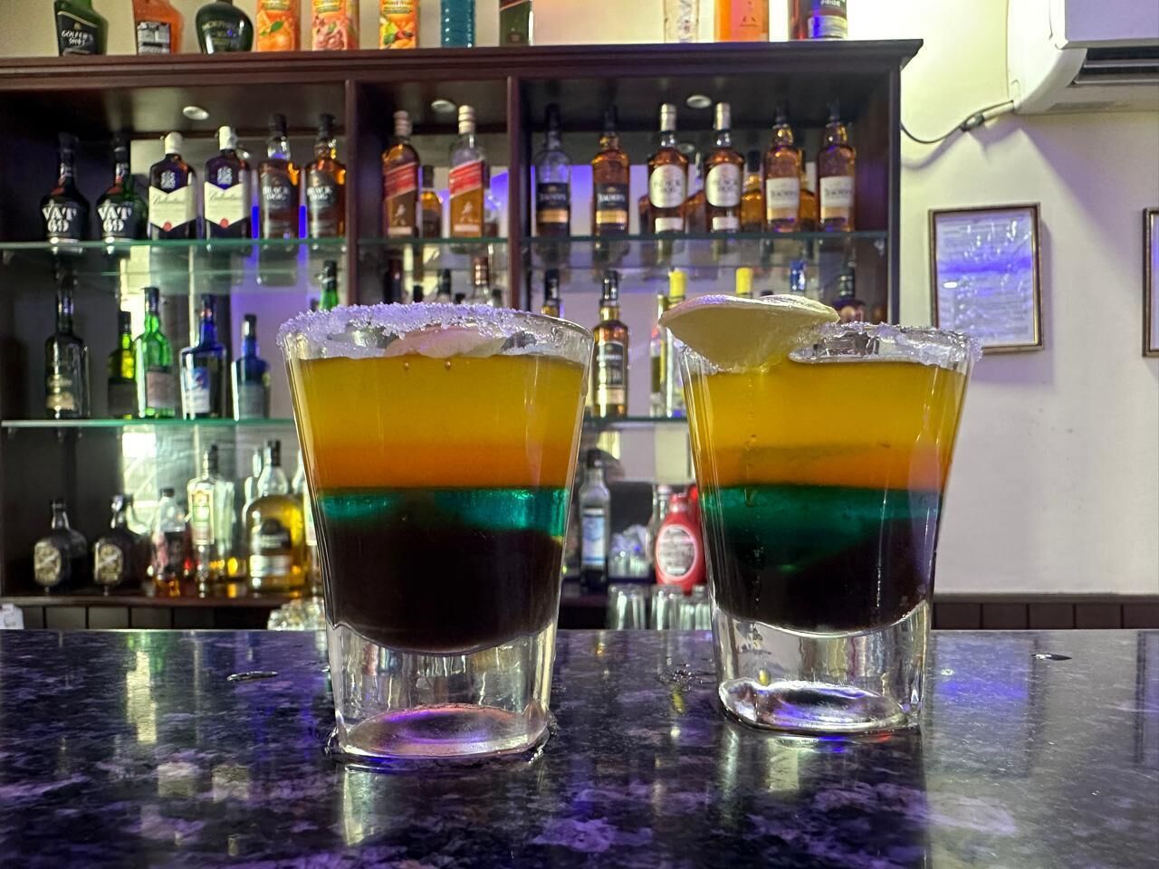 Due bicchieri di cocktail a strati su un bancone del bar, con una scaffalatura di bottiglie di liquore sullo sfondo.