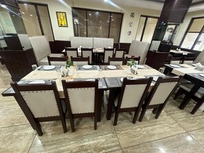 A dining area with a large rectangular table set for multiple people, surrounded by chairs with cushioned backs. - Hotel Anant Restaurant And Bar (Balh)