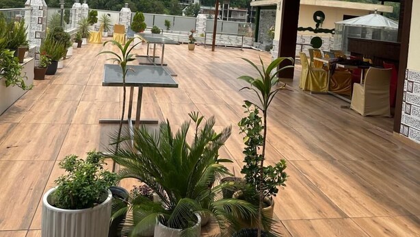 A rooftop terrace with wooden flooring, potted plants, outdoor seating, and a view of a hill with greenery and a tower in the distance. - Hotel Anant Restaurant And Bar (Balh)