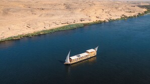 Een rivierdampboot met een opvallend zeilontwerp, die op een rivier vaart met een woestijnlandschap op de achtergrond.