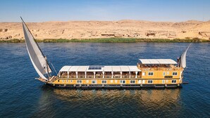 Een rivierboot met meerdere dekken en een groot zeil, drijvend op een rivier met een woest landschap op de achtergrond.