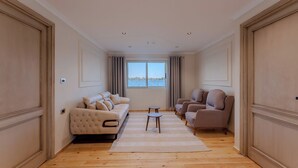 Een ruime woonkamer met een groot raam, een beige bank, een kleinere bank en twee fauteuils.