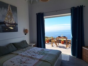 A bedroom with a bed, a nightstand, a wall-mounted lamp, and a view of the ocean through a large window with curtains. - Villa PermaVista  with excellent climate zone (Tijarafe)