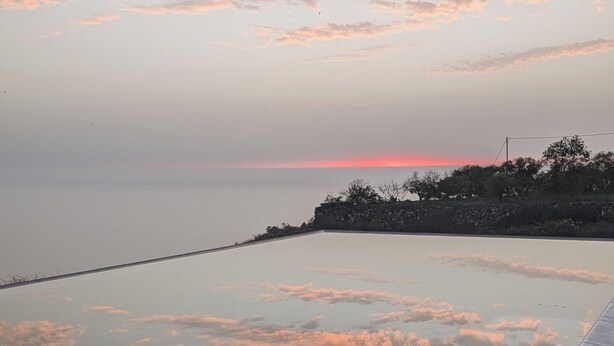 Helderblauwe lucht met wolken die zich spiegelen in een kalm wateroppervlak, een houten terras en een stenen muur begroeid met beplanting.