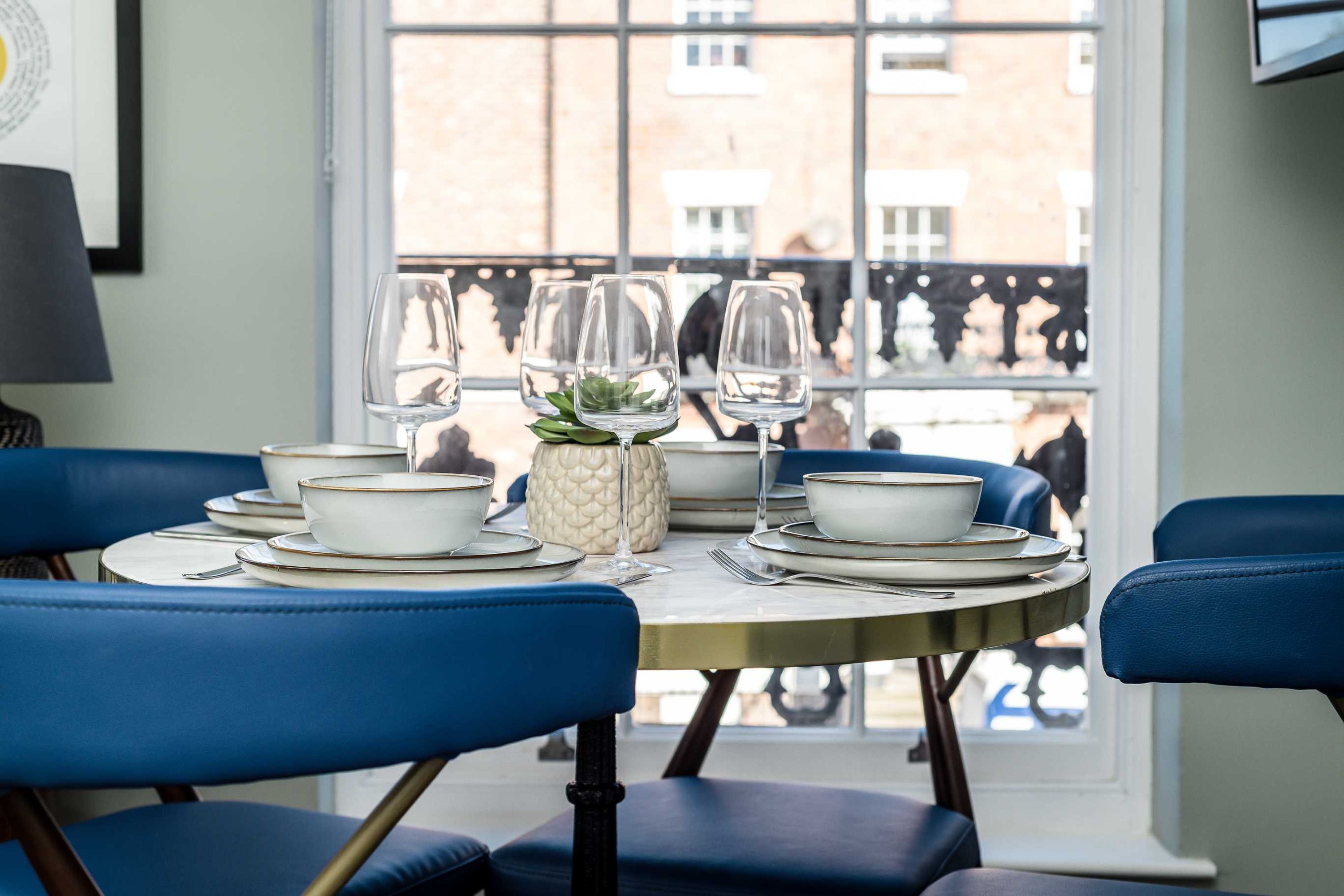 A dining table set with white plates, bowls, and glasses, surrounded by blue chairs, with a window view of buildings.