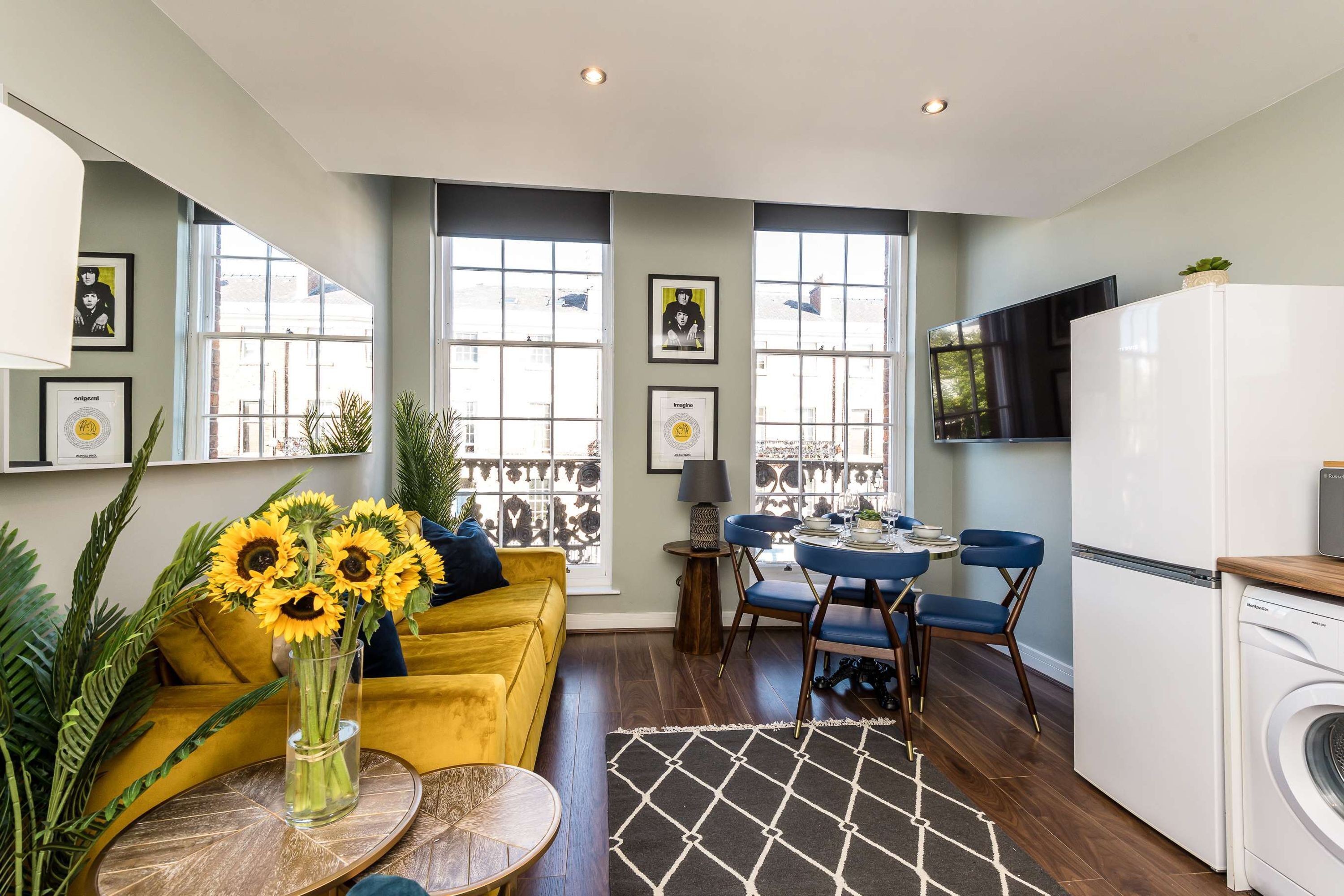 A modern living room with a yellow sofa, a dining table with blue chairs, and a kitchen area with a washing machine.