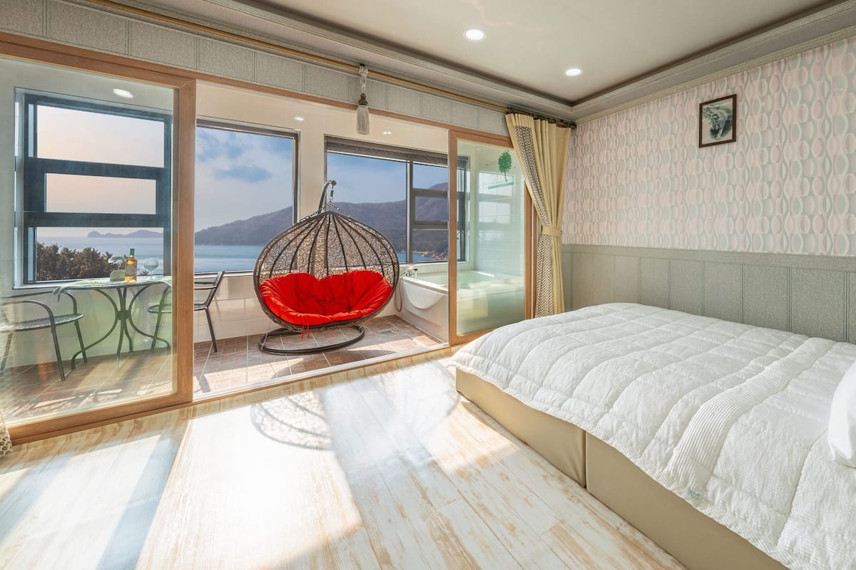 A bedroom with a large bed, a hanging chair, a bathtub, and a view of the sea.