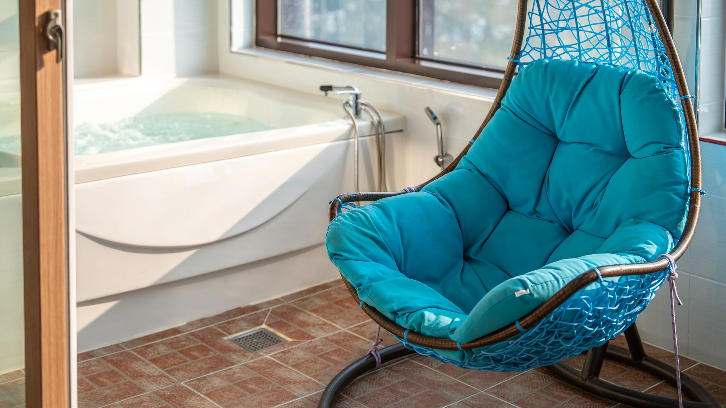 Una silla colgante turquesa en un baño con bañera y suelo de baldosas.