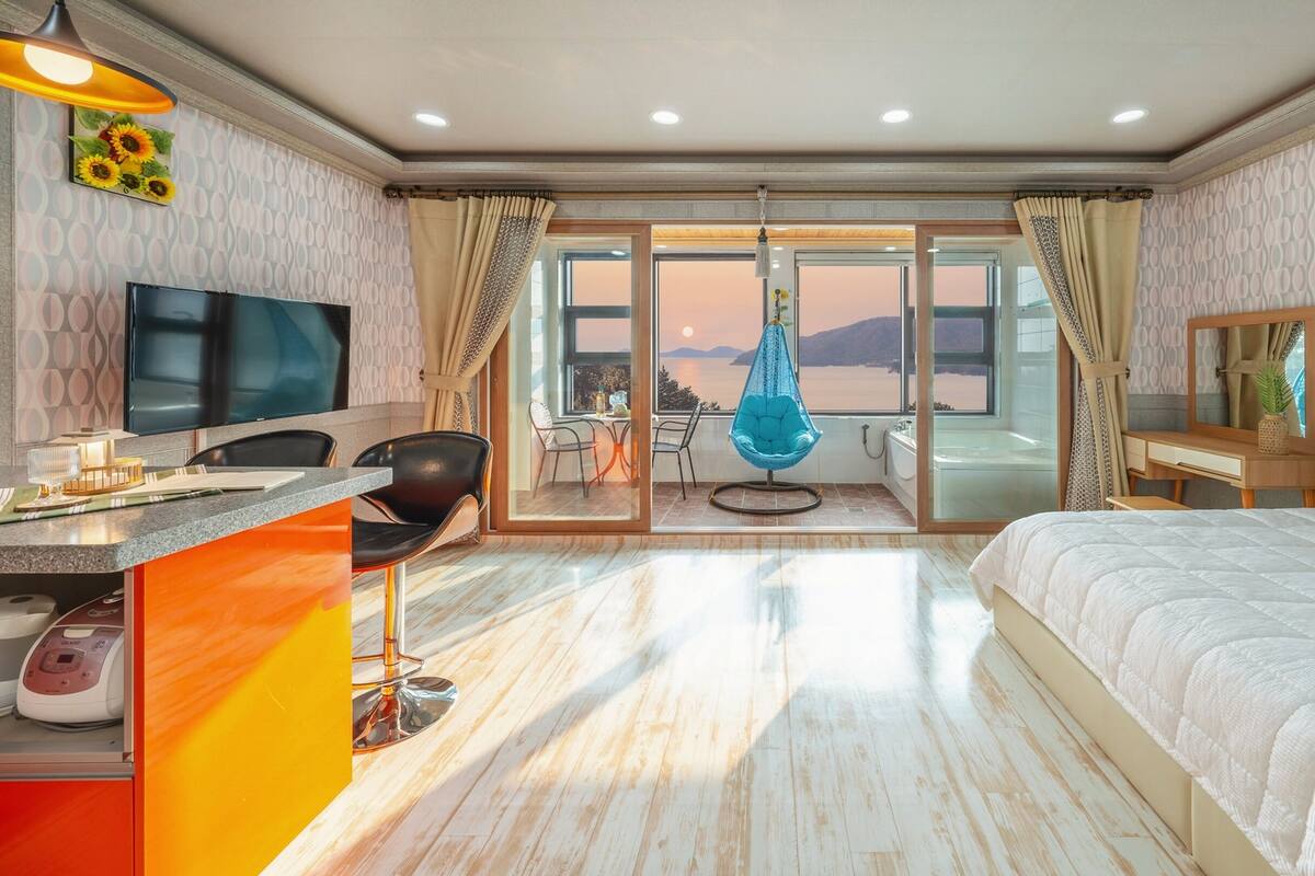 A hotel room with a bed, a desk with a chair, a TV, a massage chair, and a view of the sea.