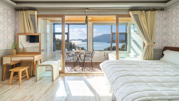 Un dormitorio con una cama grande, un escritorio, una silla, una mesita con bebidas y vistas al mar a través de amplios ventanales.