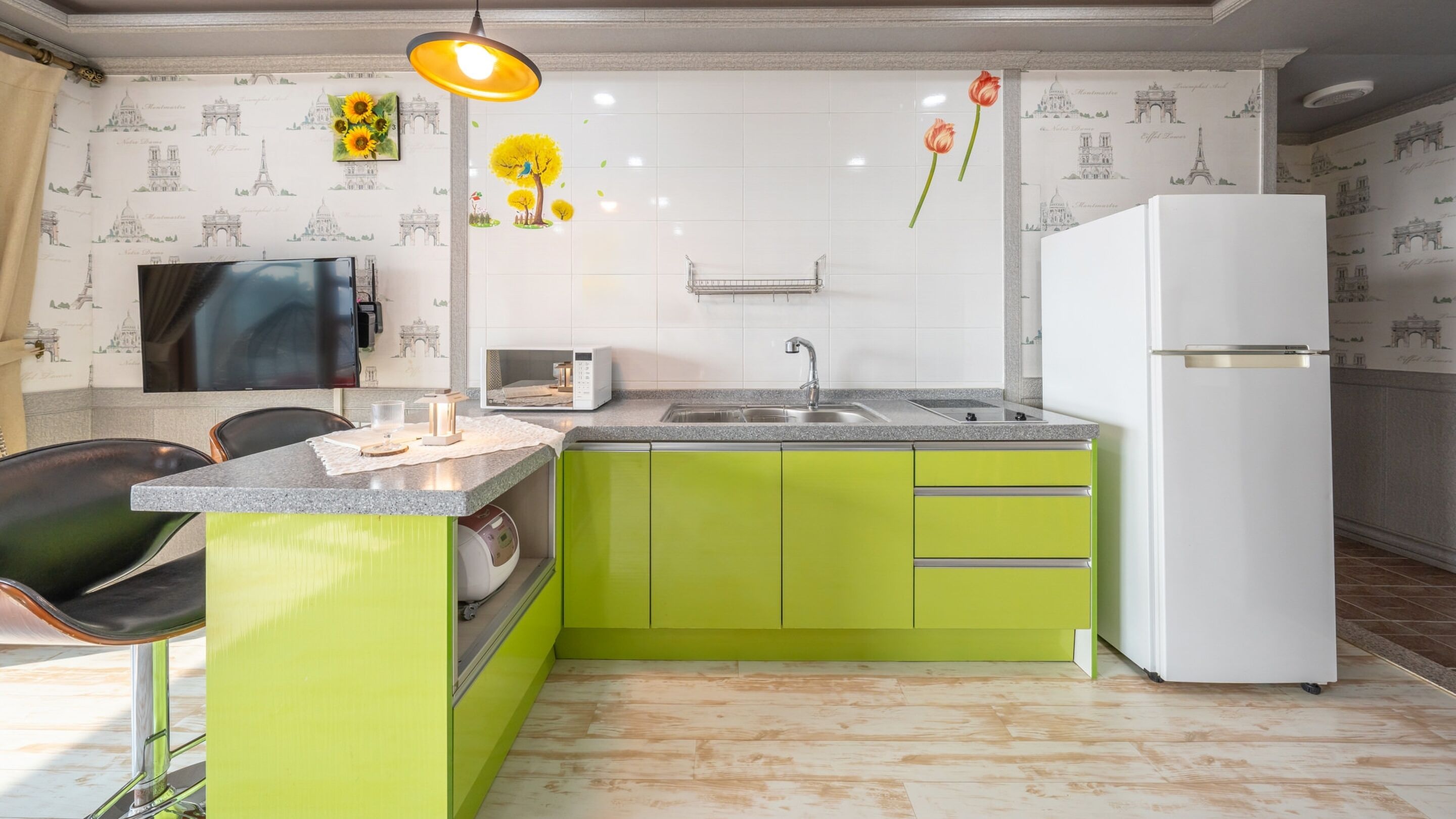 Une cuisine avec un îlot central vert, des carreaux blancs, un évier, un réfrigérateur blanc et un téléviseur fixé au mur.