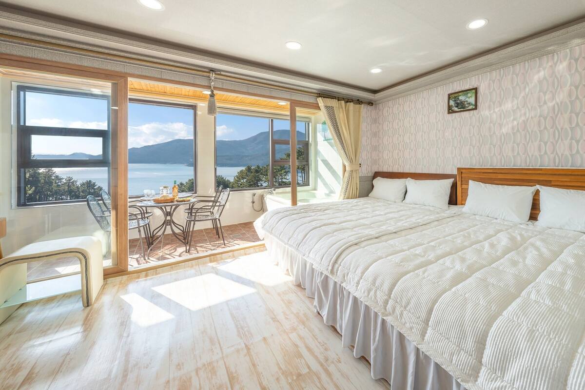 A bedroom with a large bed, a dining table, and chairs, a view of the sea, and a balcony.