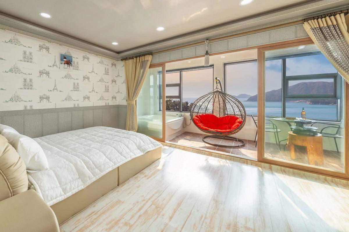 A bedroom with a bed, a hanging chair, a bathtub, and a view of the sea.